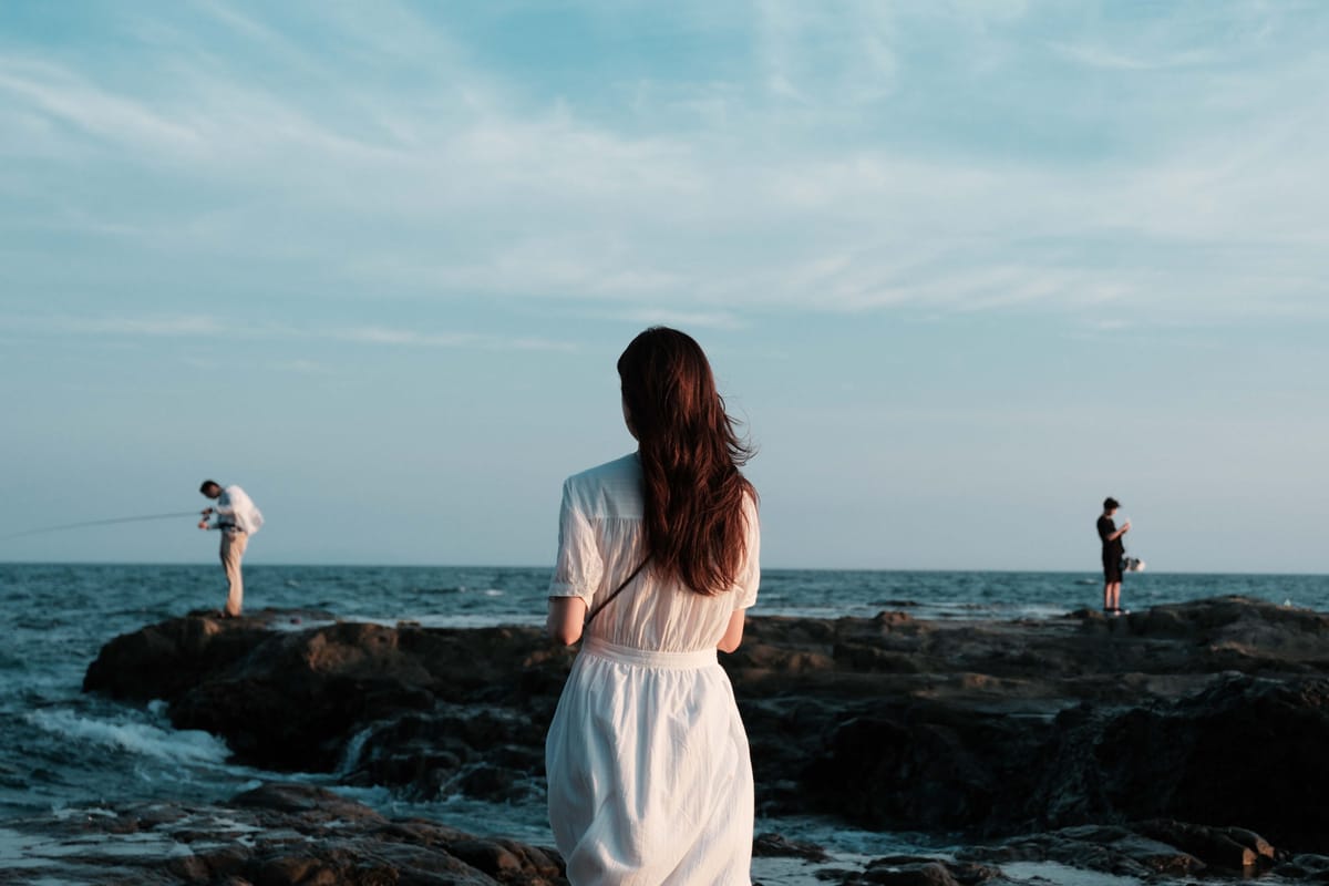 A woman standing on a rocky shore at dusk, looking out to sea