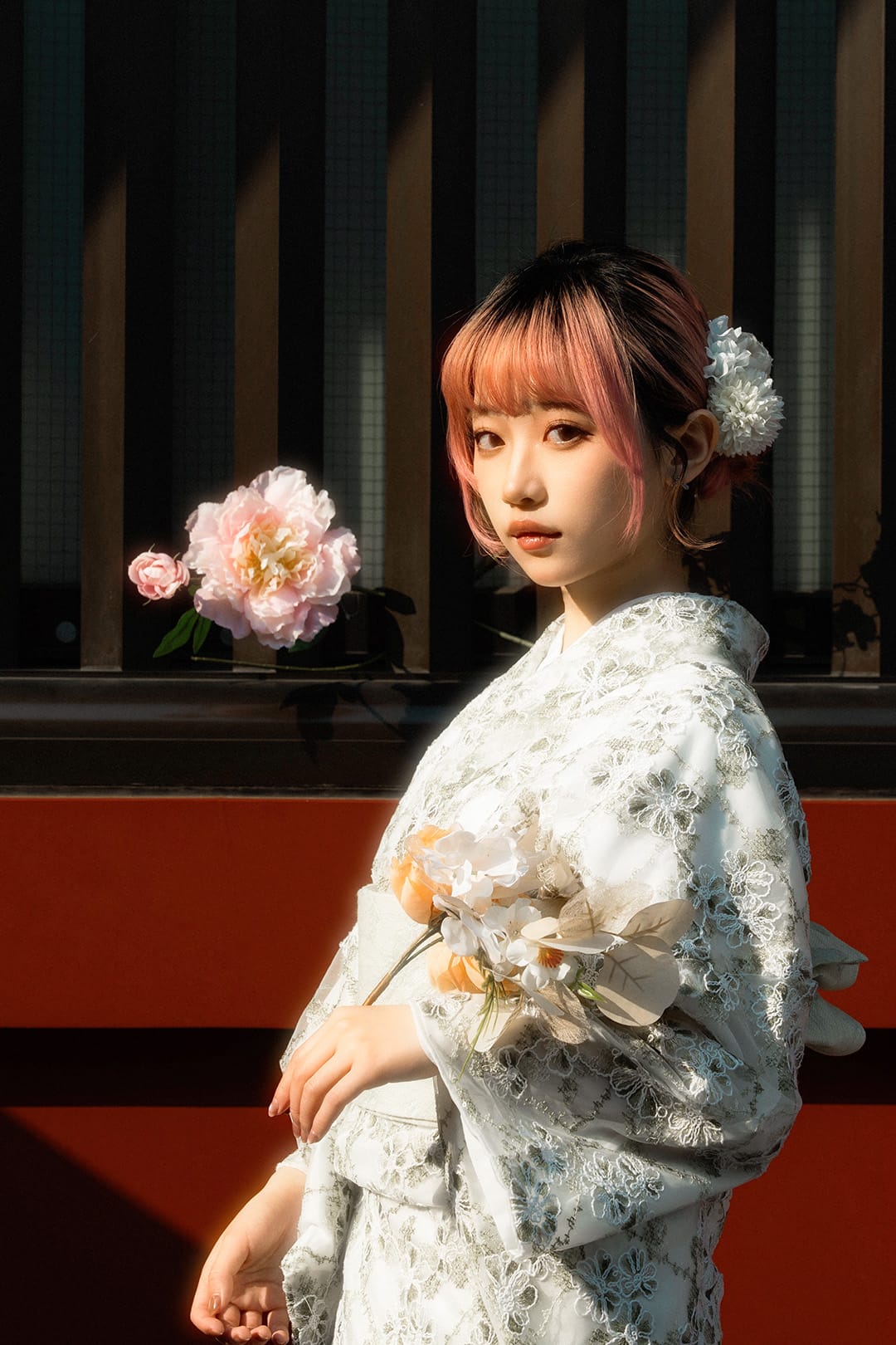 Woman in a white lace kimono with pink hair, against a red wall and hard sunlight